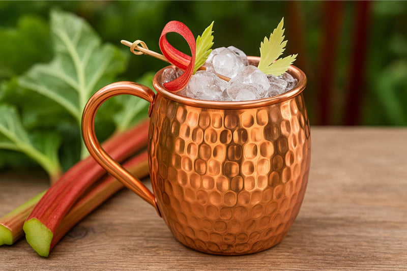 Erfrischender Rhabarber-Cocktail mit Eiswürfeln und frischer Minze, stilvoll serviert in einem glänzenden Kupferbecher auf einem Holztisch im Sonnenlicht.