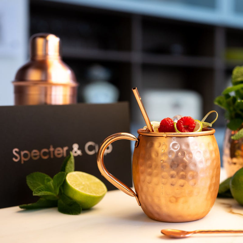 Kupferbecher Geschenk-Set von Specter & Cup. Schwarze Premiumverpackung. Auf dem Tisch steht ein Kupferbecher mit einem Kufperstrohhalm und Cocktail. Neben dem gehämmerten Kupferbecher mit Henkel liegt ein Kufperlöffel für Cocktails.